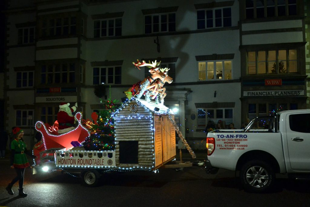 A carnival float with two reindeer and Santa in a sleigh passing the front of the Manor House in Honiton at night.