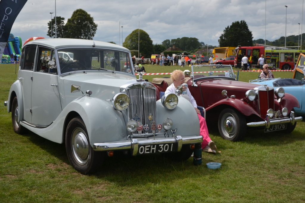 Two vintage cars and a lady sitting between them with a cup of coffee at Honiton Charter Day 2025.